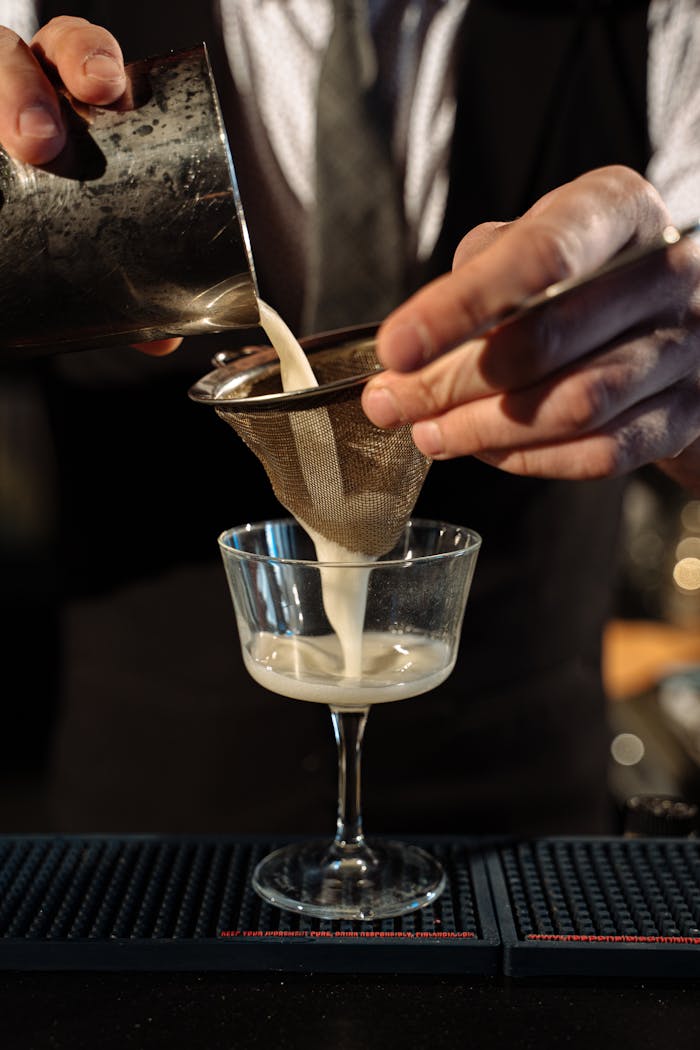 services-03 Close-up of a mixologist pouring a cocktail through a strainer into a glass at a bar.